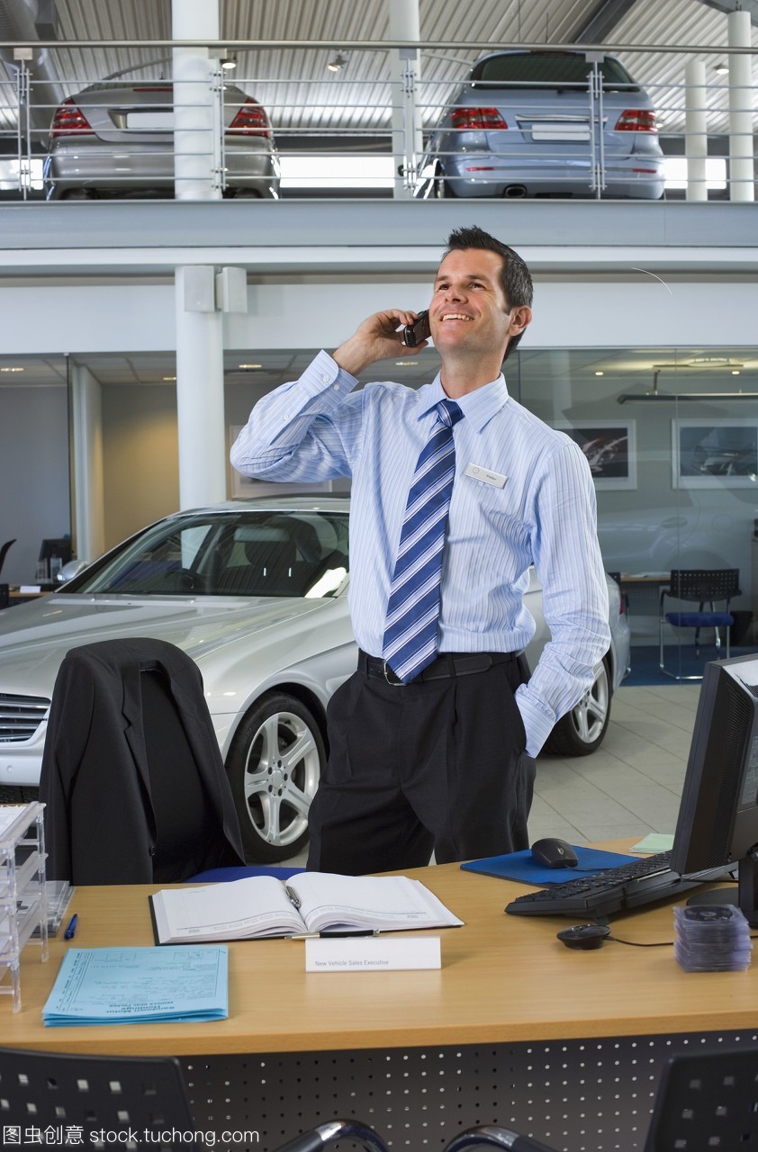 汽车销售员站在柜台旁的大型汽车陈列室里,用手机微笑着